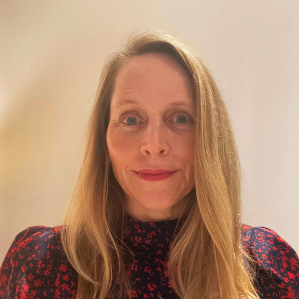 An individual with blonde hair wearing a red blouse in front of a neutral wall background.