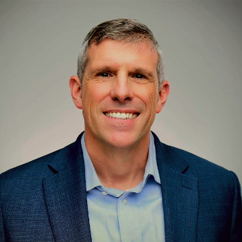An individual wearing a blue blazer and blue shirt in front of a gray background.
