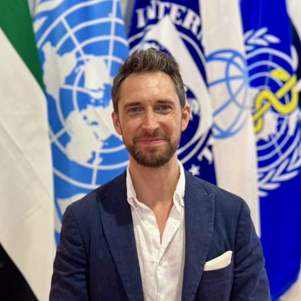An individual with short hair and a beard wearing a blue blazer and white shirt, in front of flags representing international organizations.