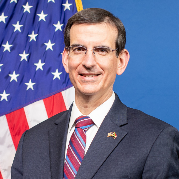 An individual wearing glasses, a gray suit, and red striped tie posing in front of an American flag.
