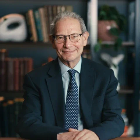 A man with glasses, grey hair, and a dark blue suit with a blue necktie.