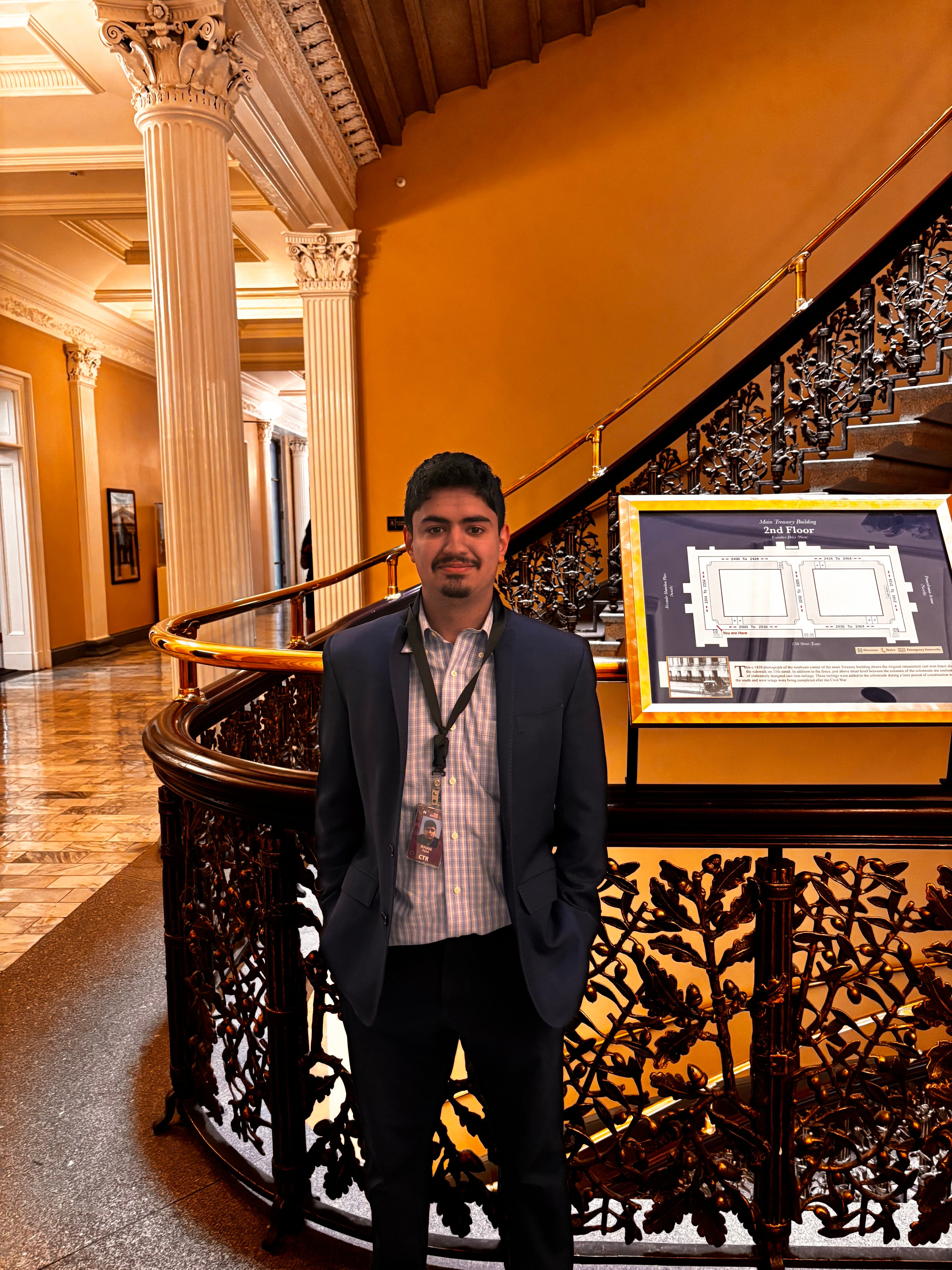 A person stands in a building with elegant architecture, featuring ornate railings and tall columns. A display screen with floor plans is visible nearby.