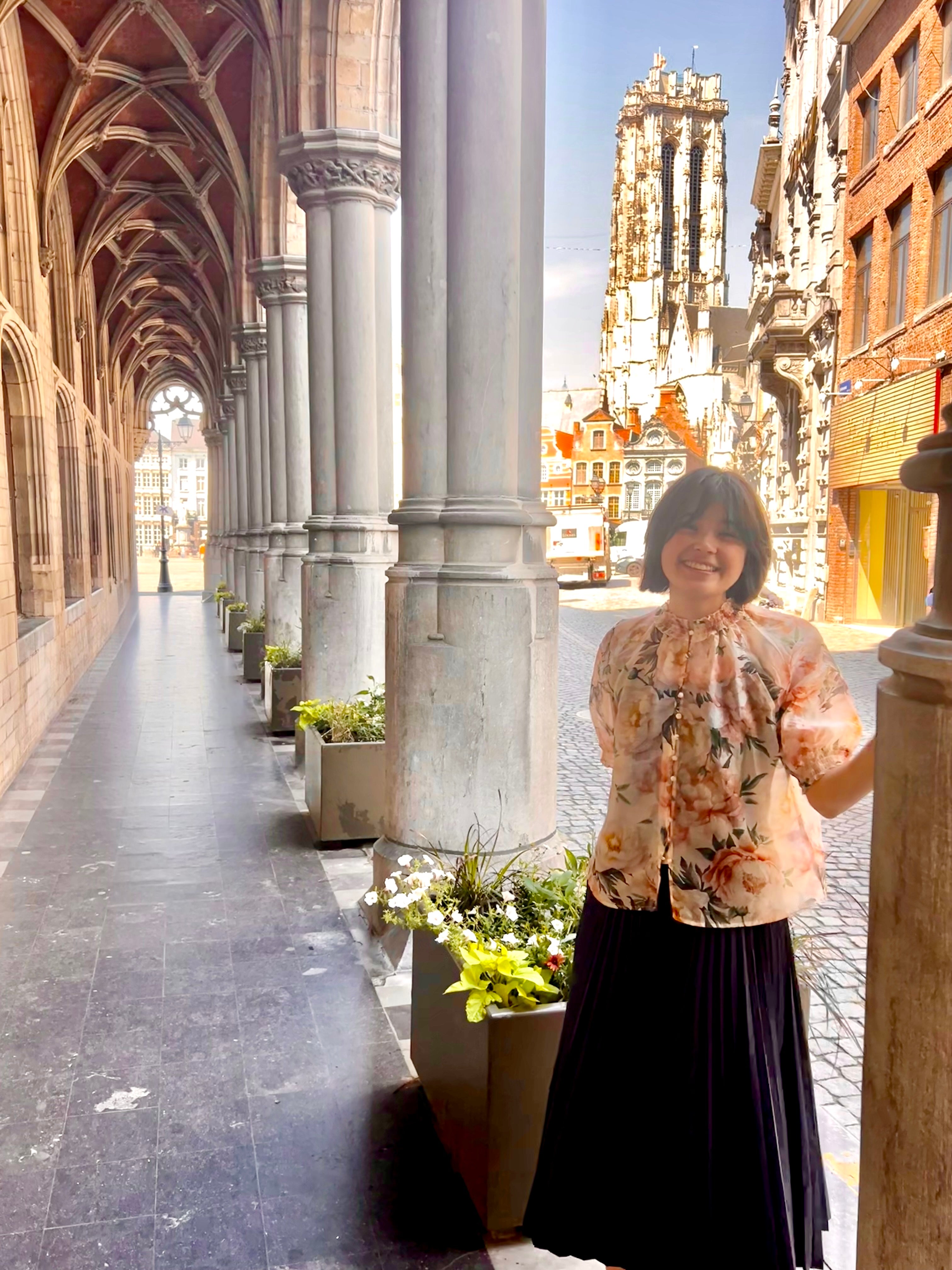 A person stands smiling in a decorated corridor with tall arches and columns adorned with flower planters.