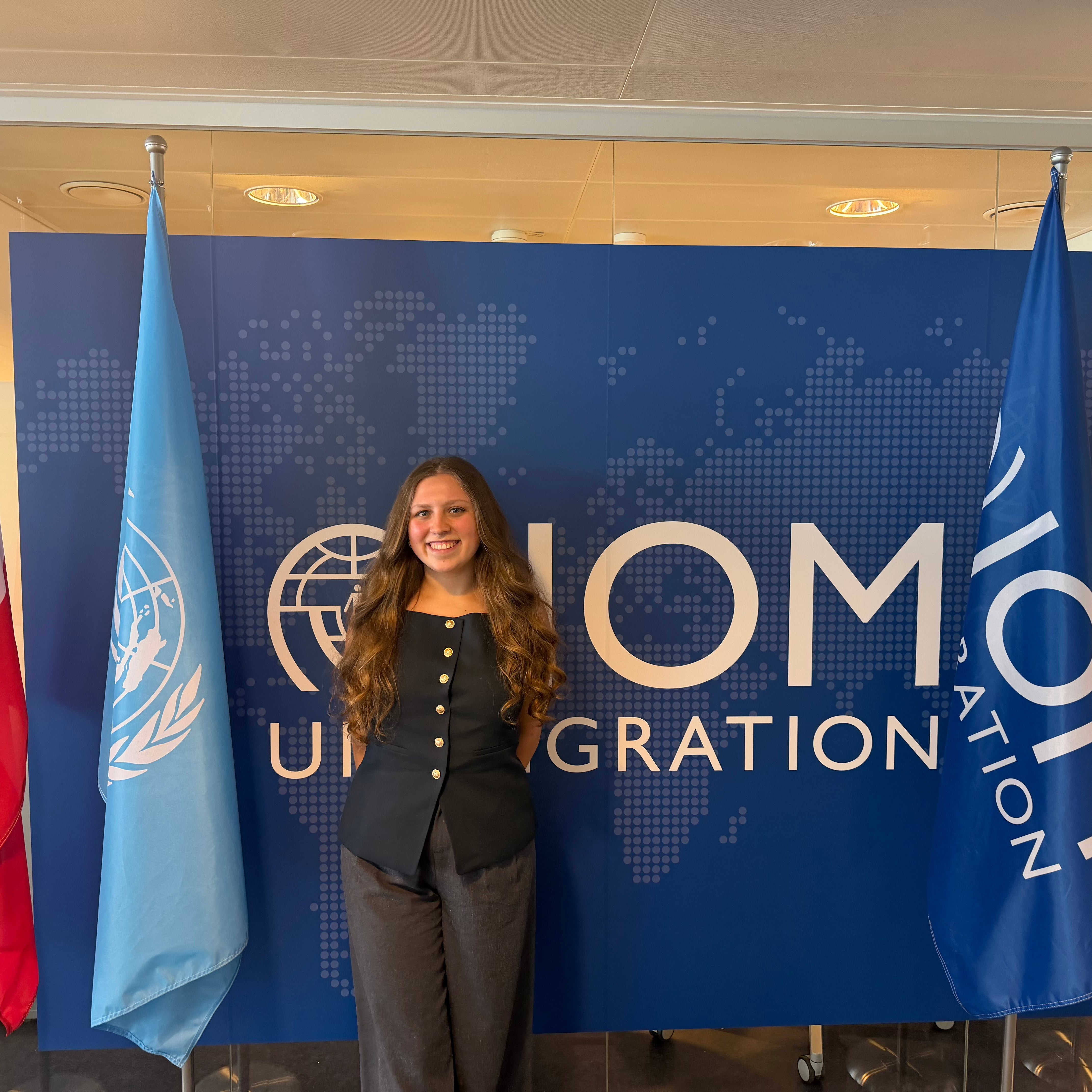 A person stands smiling in front of a blue wall with the International Organization for Migration (IOM) logo. The background features a world map design.