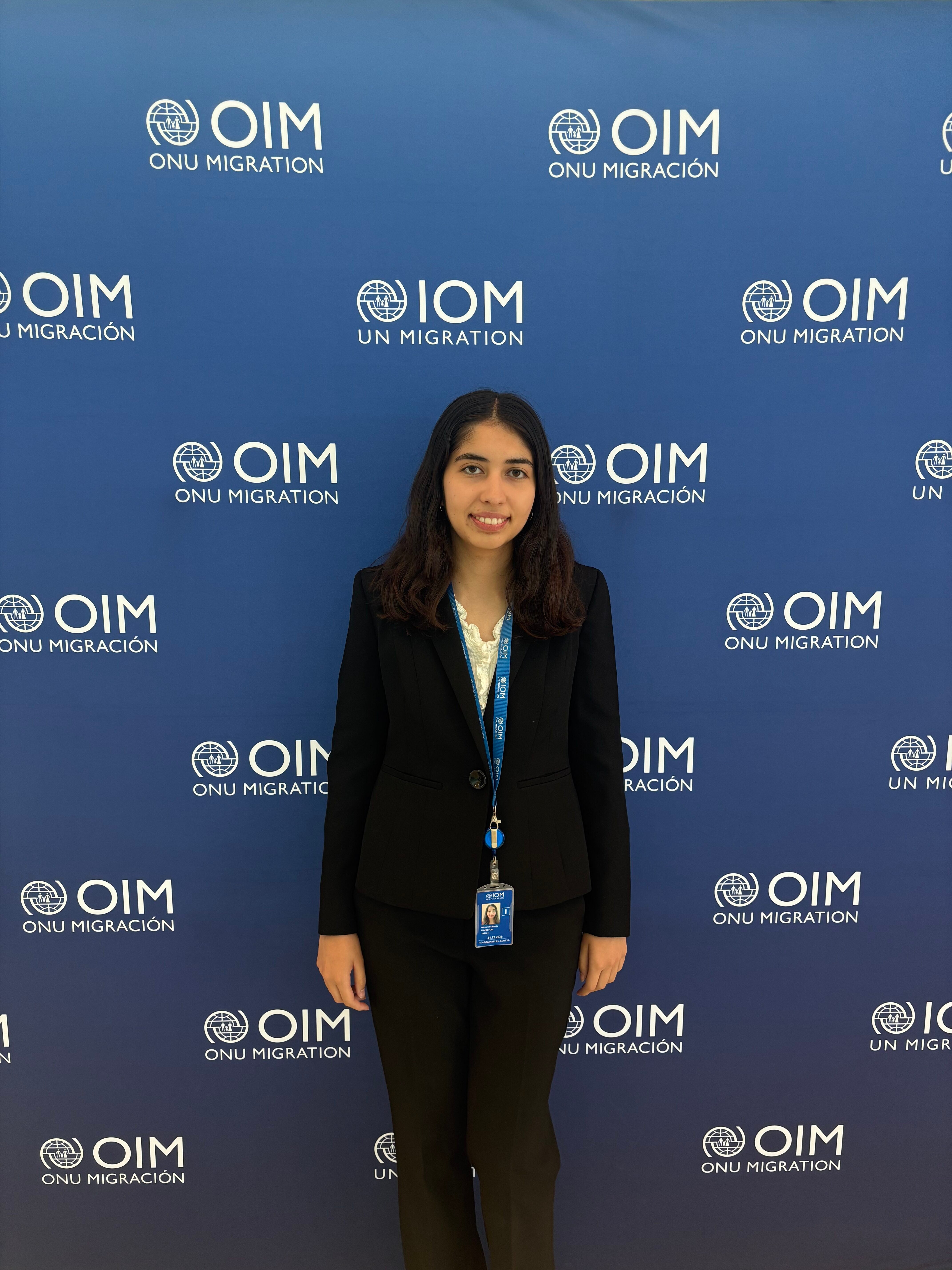 A person stands in front of a blue backdrop with the OIM ONU Migration logo repeatedly printed on it.