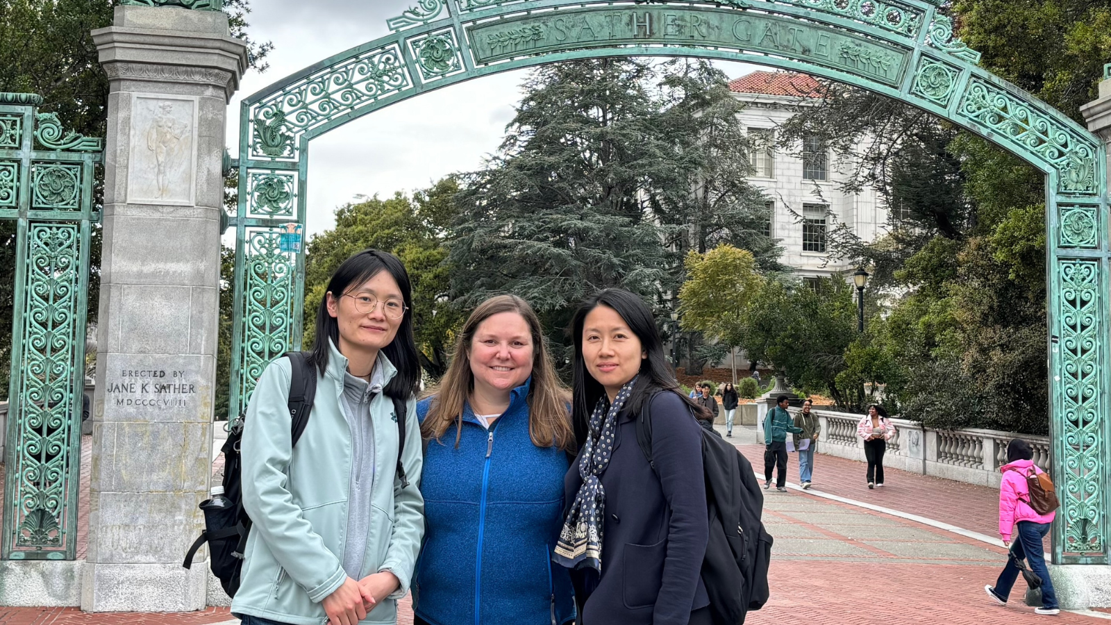 Three individuals stand smiling in front of the iconic Sather Gate at the University of California, Berkeley. The sky is overcast, and a few people stroll in the background.