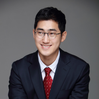 A man with short black hair, wearing glasses and a suit posing in front of a gray background.