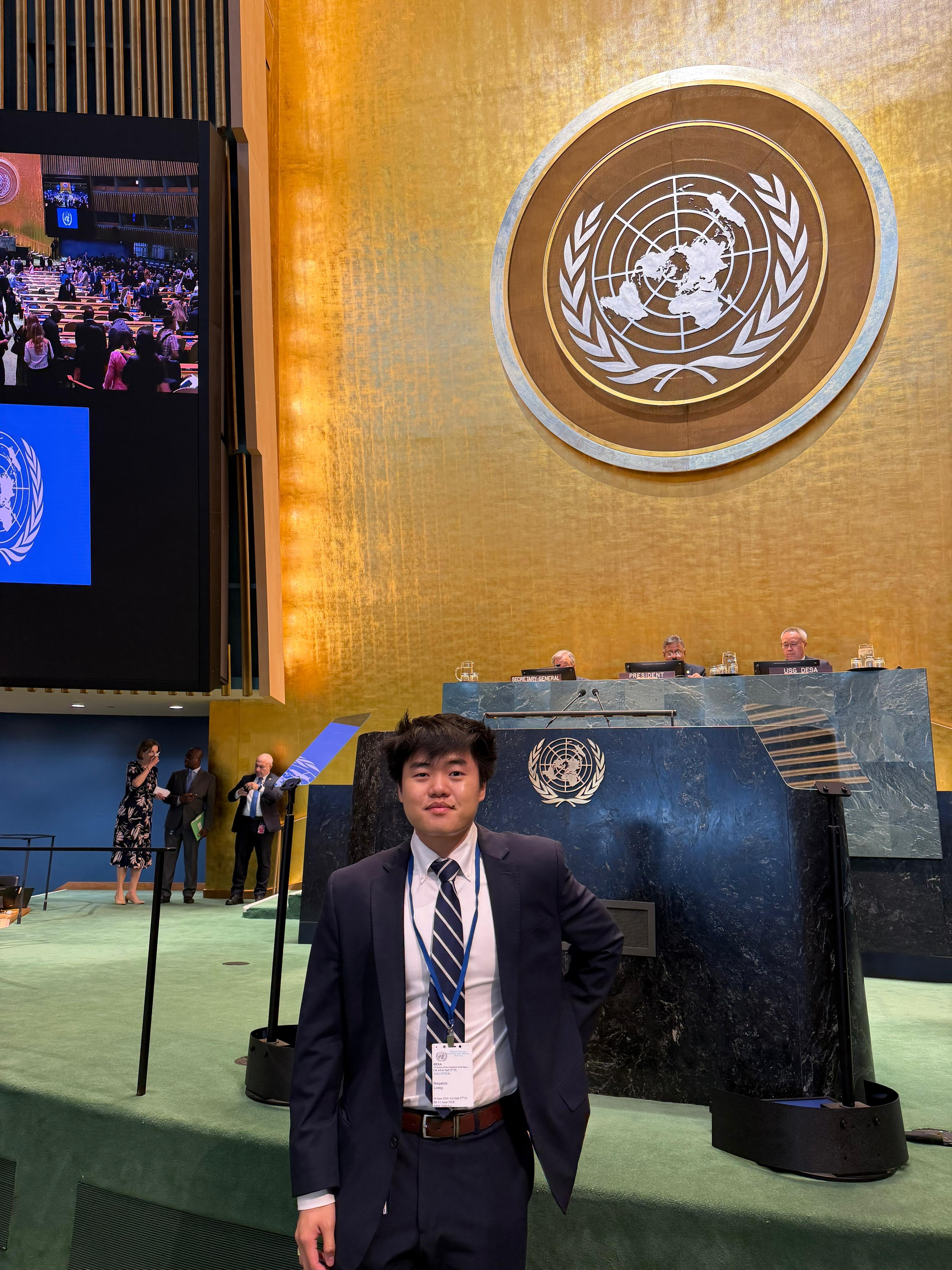 A person stands in a formal suit inside the United Nations General Assembly Hall. The UN emblem is prominently displayed on the golden wall behind them.