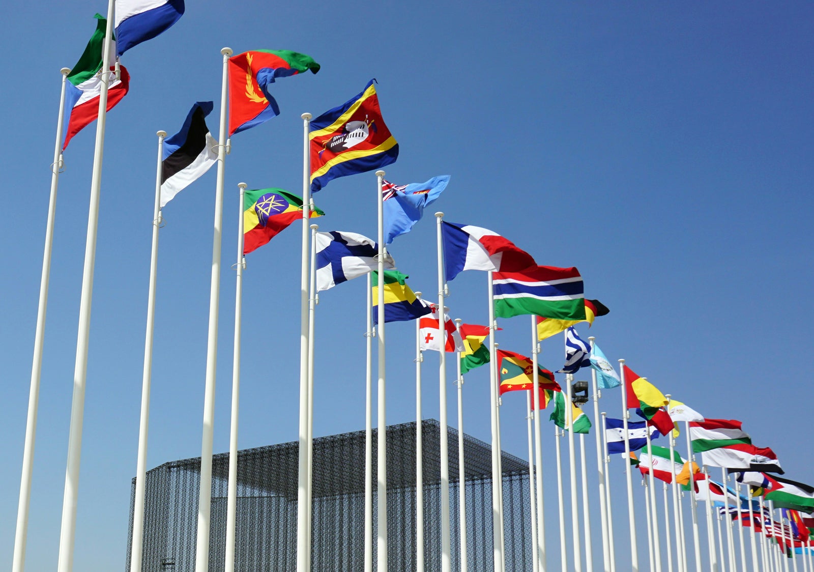 A group of international flags, including those of France and the United States, fluttering in the wind against a clear blue sky. The flags are mounted on white poles, and a modern building is visible in the background.