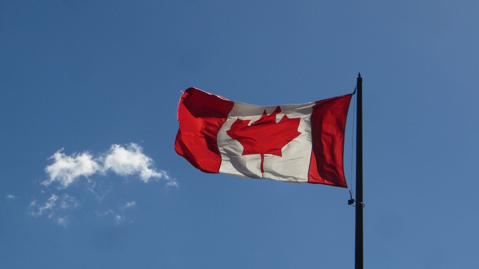 The image shows the Canadian flag, featuring a red maple leaf on a white background with red borders, flying against a clear blue sky with a small white cloud.