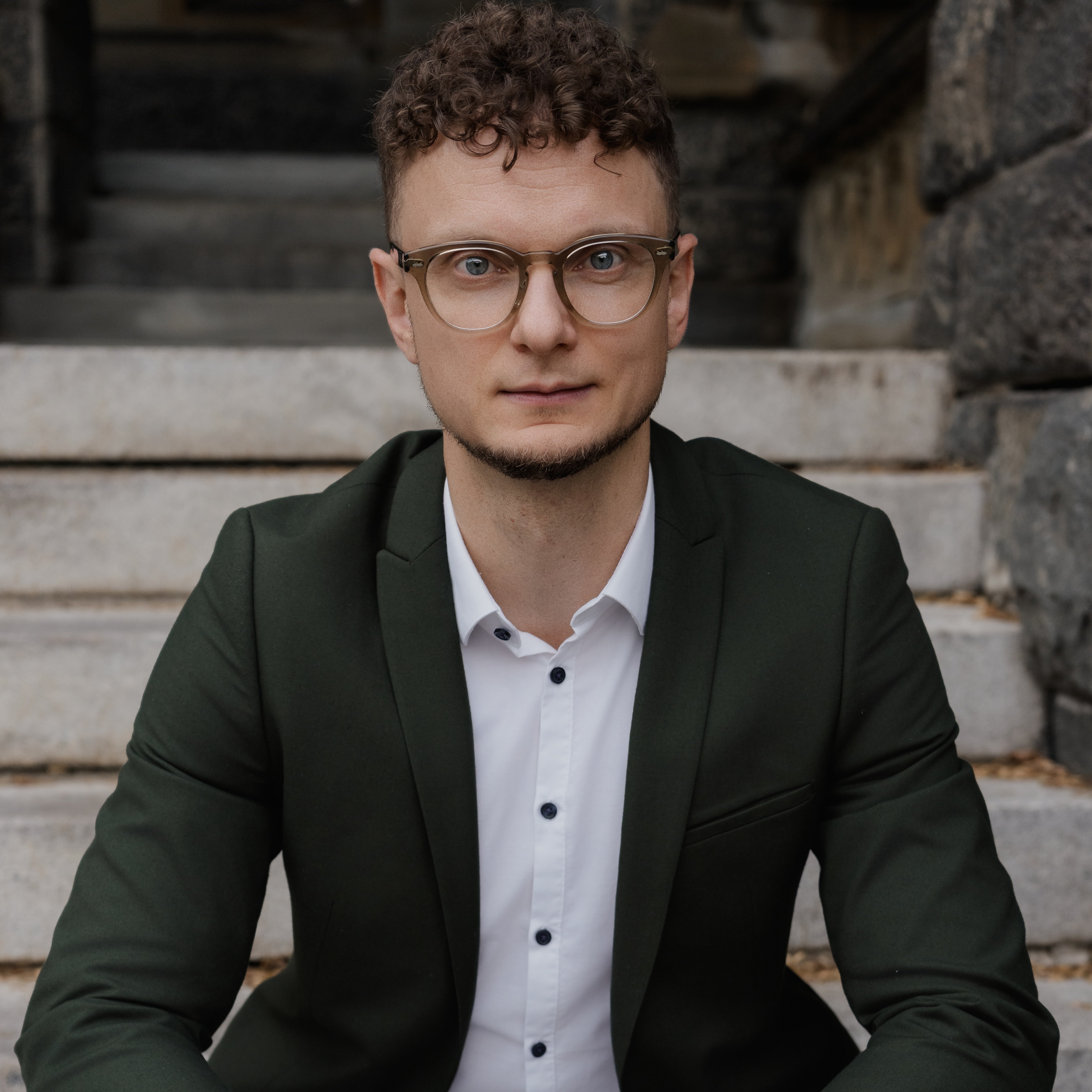 A man with short curly brown hair wearing glasses and a green blazer, sitting on the steps of Georgetown's Healy Hall.