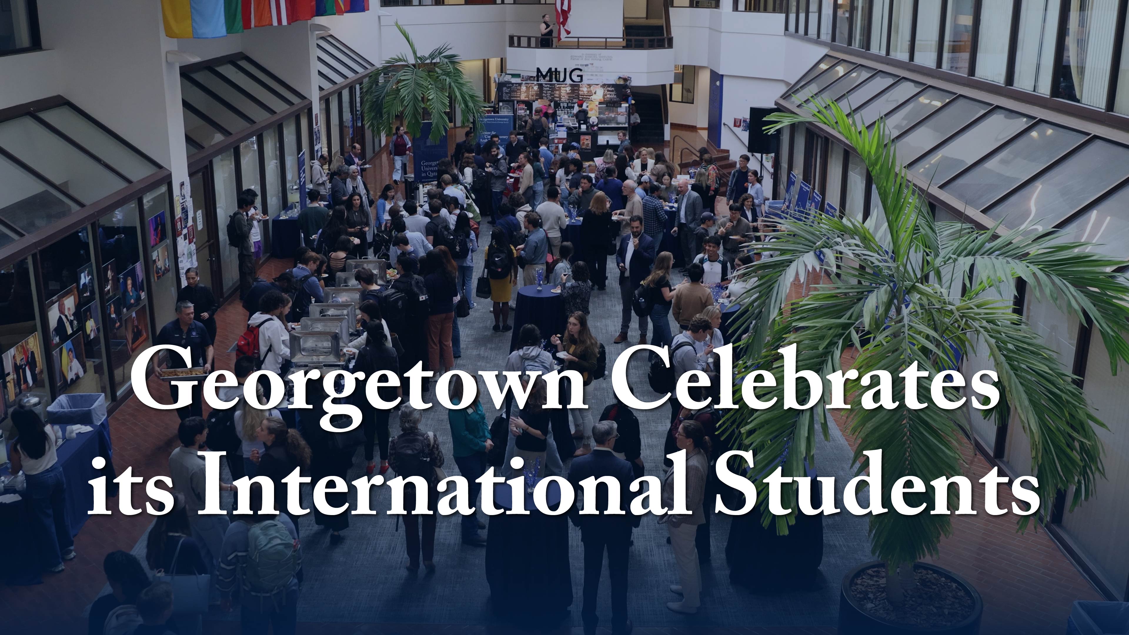 A large group of people gathers in a spacious, bright indoor area at Georgetown University. The space is decorated with international flags, and there are tall windows and palm plants. The text overlay reads, "Georgetown Celebrates its International Students.