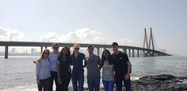 Group of college students pose in front of a bridge at midday.